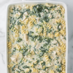 A white dish filled with creamy spinach artichoke chicken pasta topped with a sprinkle of herbs. Text reads "Spinach Artichoke Chicken Pasta - Quick Dinner Idea." A website URL is at the bottom.