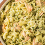 A bowl of pasta tossed with pieces of salmon and pesto sauce. Text overlay at the top reads "Pesto Salmon Pasta - Kid-Friendly." At the bottom, a banner says "Full Recipe at cookingupmemories.com.