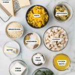 Top-down view of ingredients on a marble surface: parmesan cheese, white sharp cheddar cheese, milk, pasta, artichokes, breadcrumbs, seasonings, diced chicken, butter, sour cream, chicken broth, cream cheese, spinach, and flour.