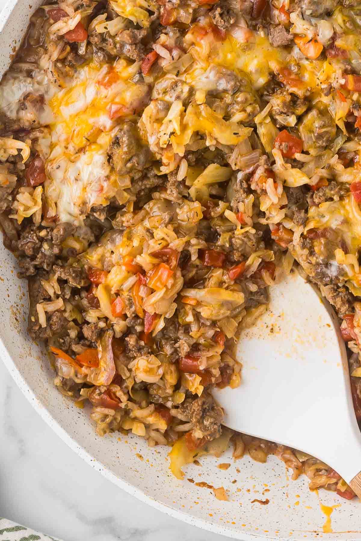 A close-up of a cooked dish featuring melted cheese, ground beef, rice, and chopped vegetables. A white spatula rests in the mixture, suggesting serving or mixing. 