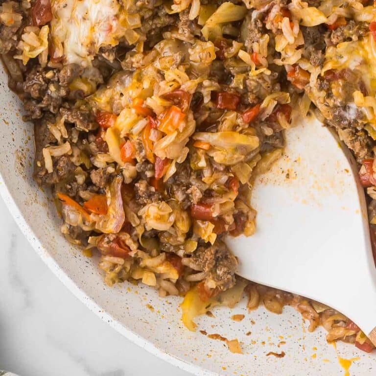 A skillet filled with cooked rice, ground meat, and tomatoes. A white spatula is partially visible, mixing the ingredients. The dish appears savory and is seasoned with various herbs and spices.