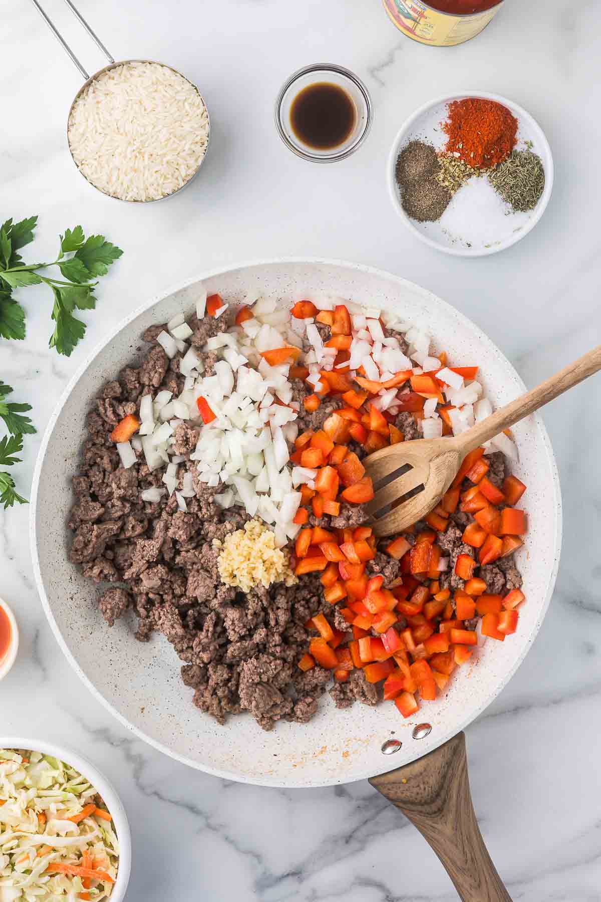 A skillet contains ground beef, chopped onions, minced garlic, and diced red bell peppers. 