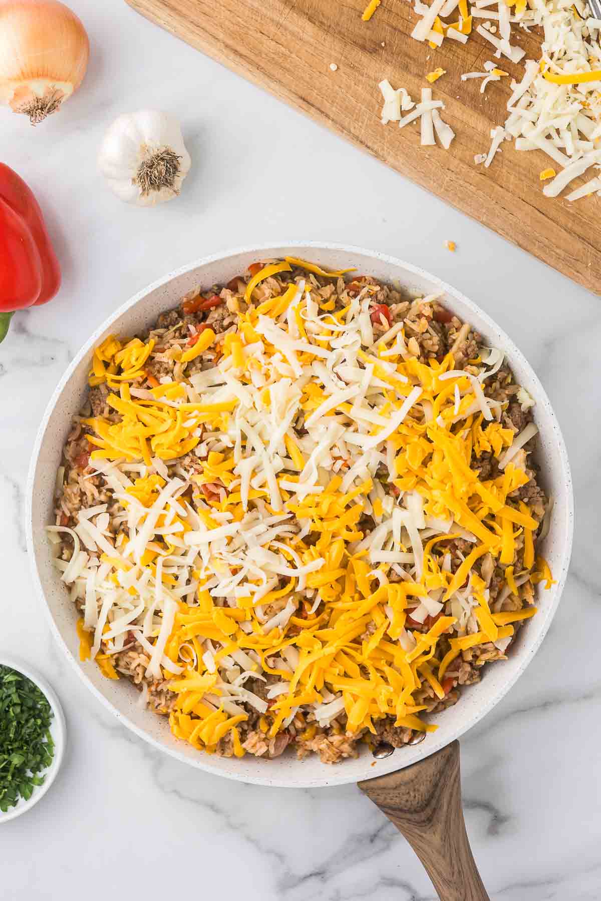 A white skillet filled with a mixture of rice, vegetables, and ground meat, topped with shredded cheddar and white cheese. 