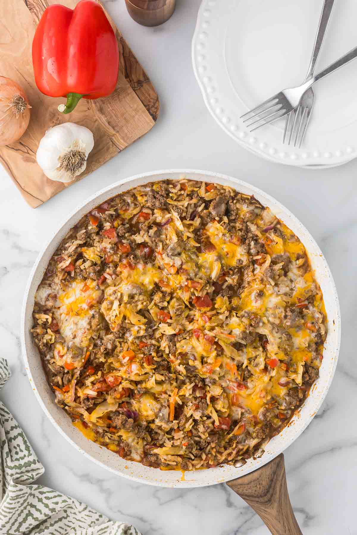 A skillet filled with a cheesy beef and rice casserole sits on a marble countertop. Nearby, a wooden board holds a red bell pepper, onion, and garlic. Two empty white plates with forks rest nearby. A green patterned cloth is in the corner.