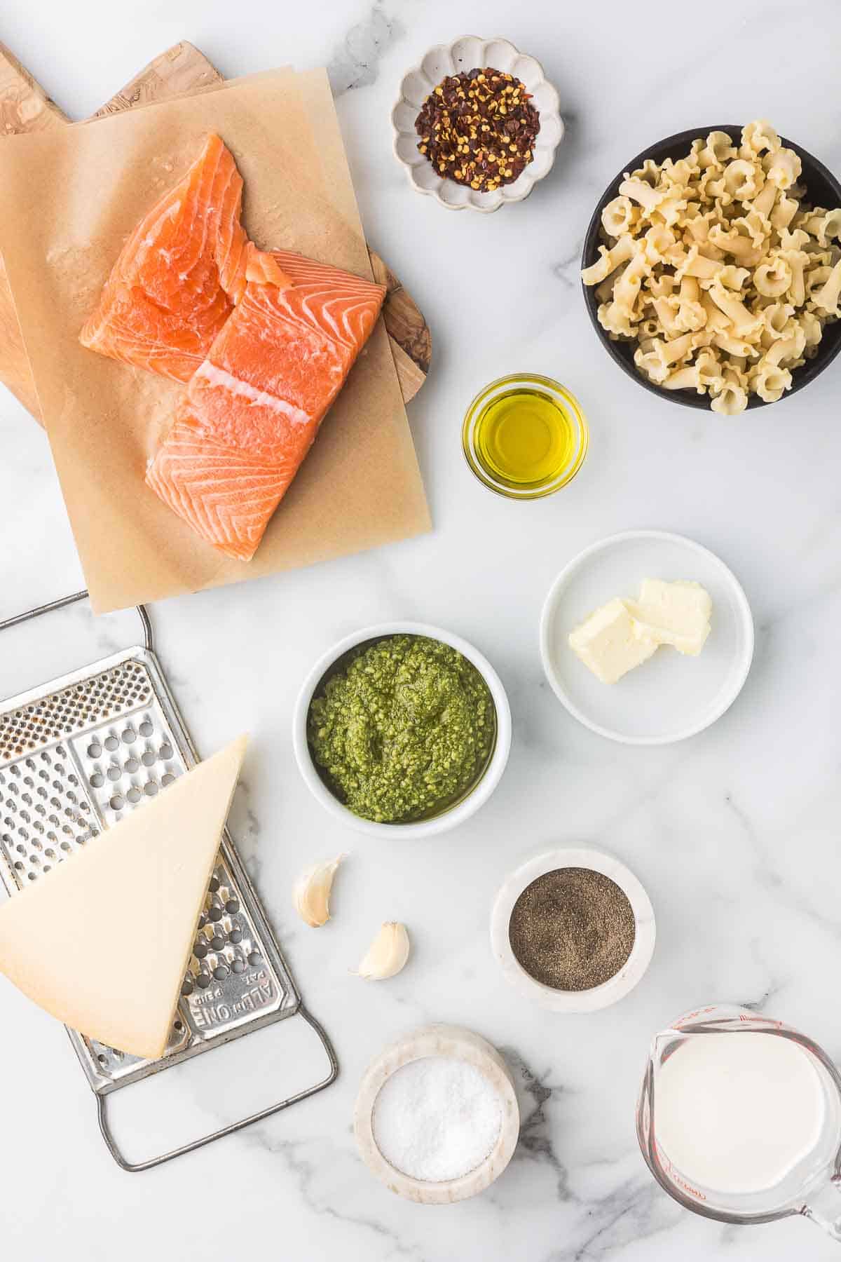 Ingredients for a salmon pasta dish are arranged on a marble surface. Visible items include raw salmon, uncooked pasta, pesto, Parmesan cheese, olive oil, butter, garlic, pepper, salt, and cream, along with red pepper flakes for seasoning.