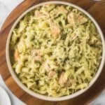 A bowl of pasta coated in a creamy pesto sauce, mixed with chunks of salmon, garnished with chopped parsley, sitting on a wooden board.