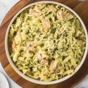 A bowl of pasta coated in a creamy pesto sauce, mixed with chunks of salmon, garnished with chopped parsley, sitting on a wooden board.