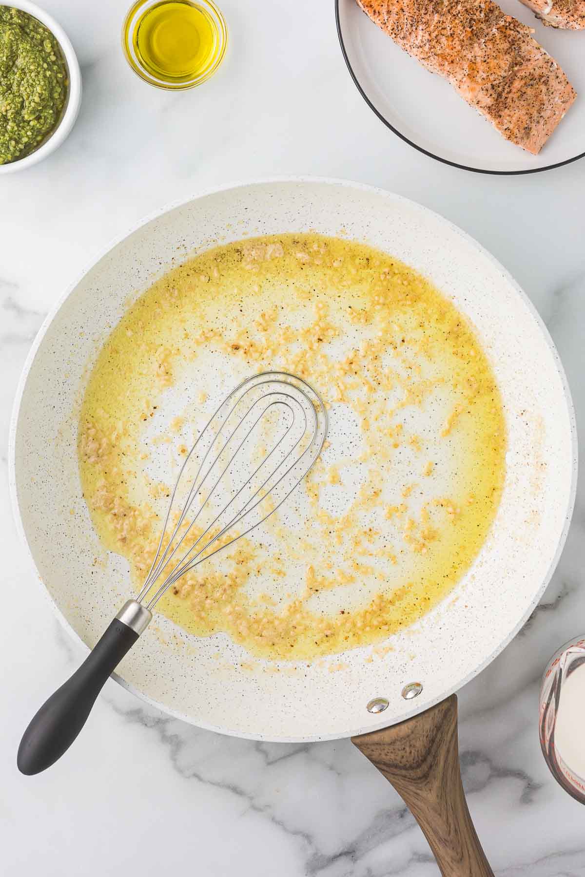 A skillet on a marble countertop containing sautéed garlic and butter with a whisk. Nearby are a plate with seasoned salmon, a bowl of pesto, a small dish of olive oil, and a jug of cream.