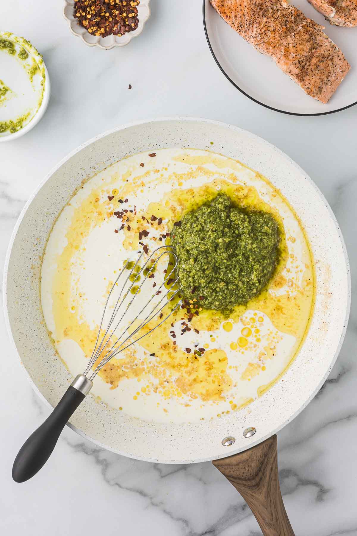 A white pan on a marble surface contains cream being mixed with green pesto sauce and red pepper flakes using a whisk. In the background, raw seasoned salmon is placed on a plate, and a small bowl contains additional red pepper flakes.