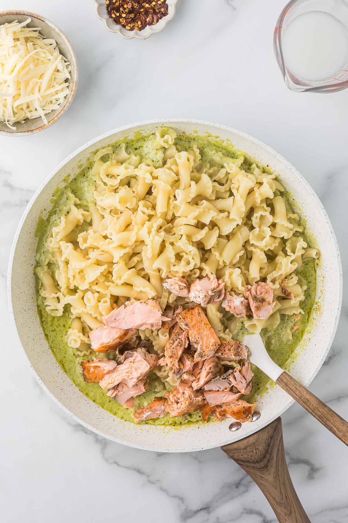 A skillet with cooked pasta in a green sauce, topped with chunks of salmon. A wooden spoon is stirring the mixture. Nearby are bowls of grated cheese, red pepper flakes, and a small pitcher of cream on a marble countertop.