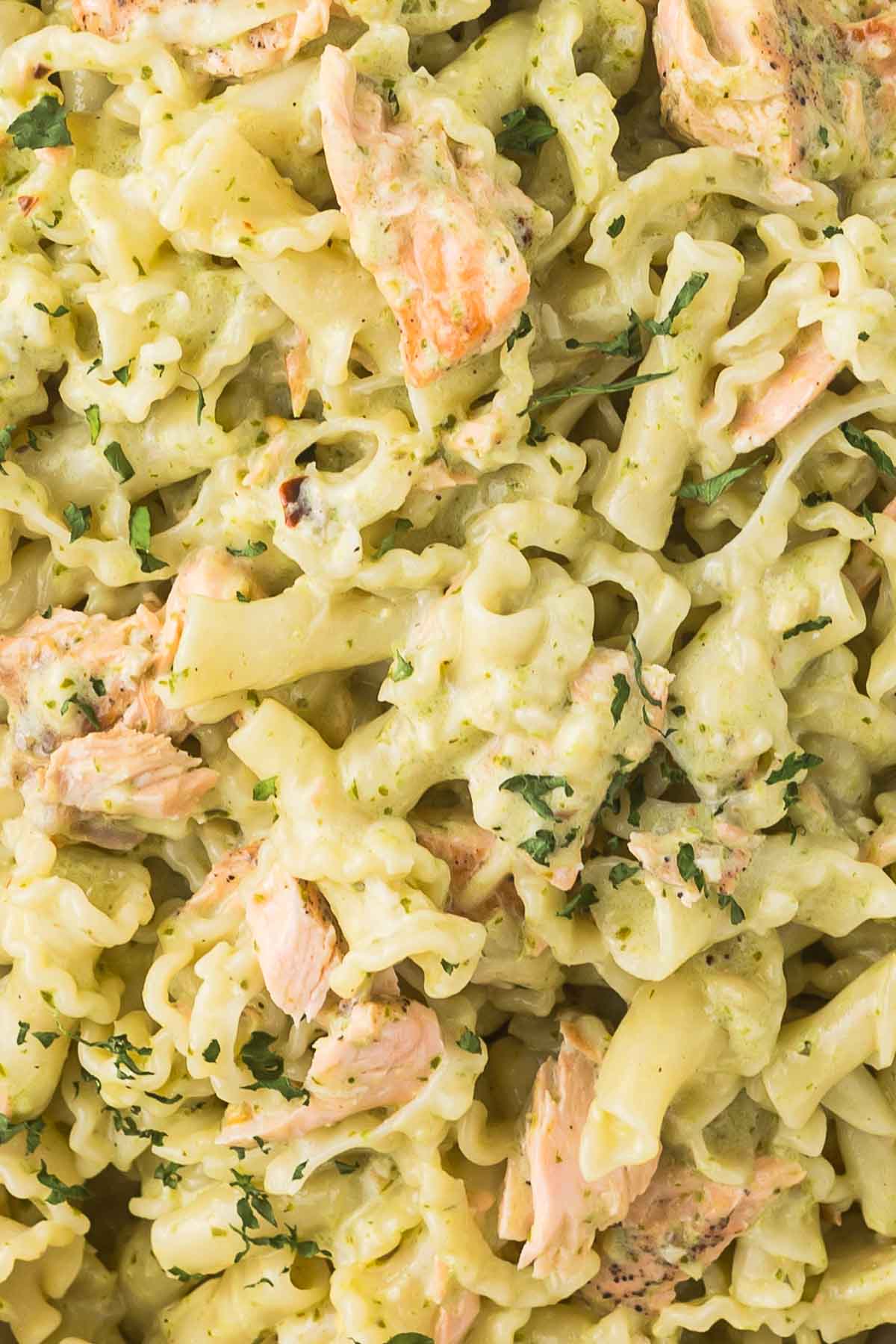 Close-up of pasta with creamy avocado sauce and chunks of salmon, garnished with chopped herbs. The dish appears rich and flavorful, with a mix of textures from the pasta and fish.