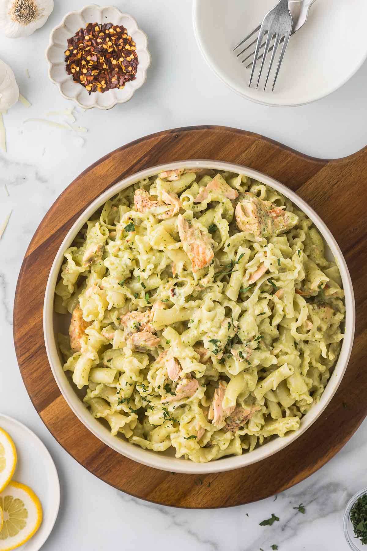 A bowl of creamy pasta with pieces of salmon and herbs, placed on a wooden serving board. Surrounding the bowl are lemon slices, a small dish of crushed red pepper, garlic cloves, and an empty plate with forks.