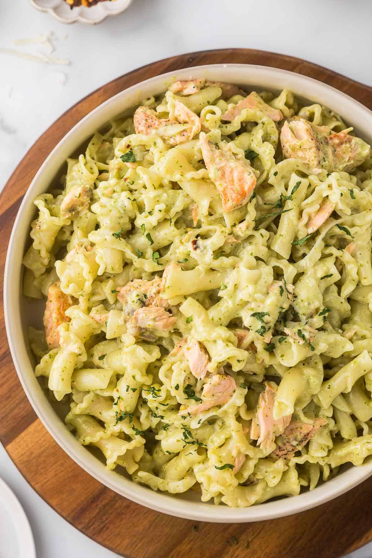 A bowl of creamy pasta with chunks of salmon and a sprinkle of fresh herbs, served on a wooden board.