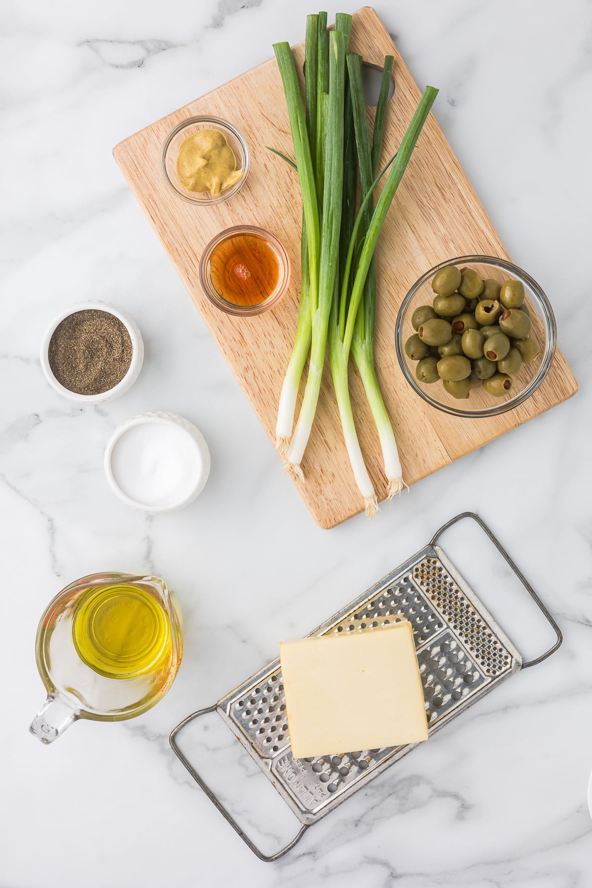 A wooden cutting board with green onions, small bowls of mustard, honey, and green olives. Next to the board are bowls of salt and pepper, a glass cup of olive oil, and a cheese block on a grater, all on a marble surface.