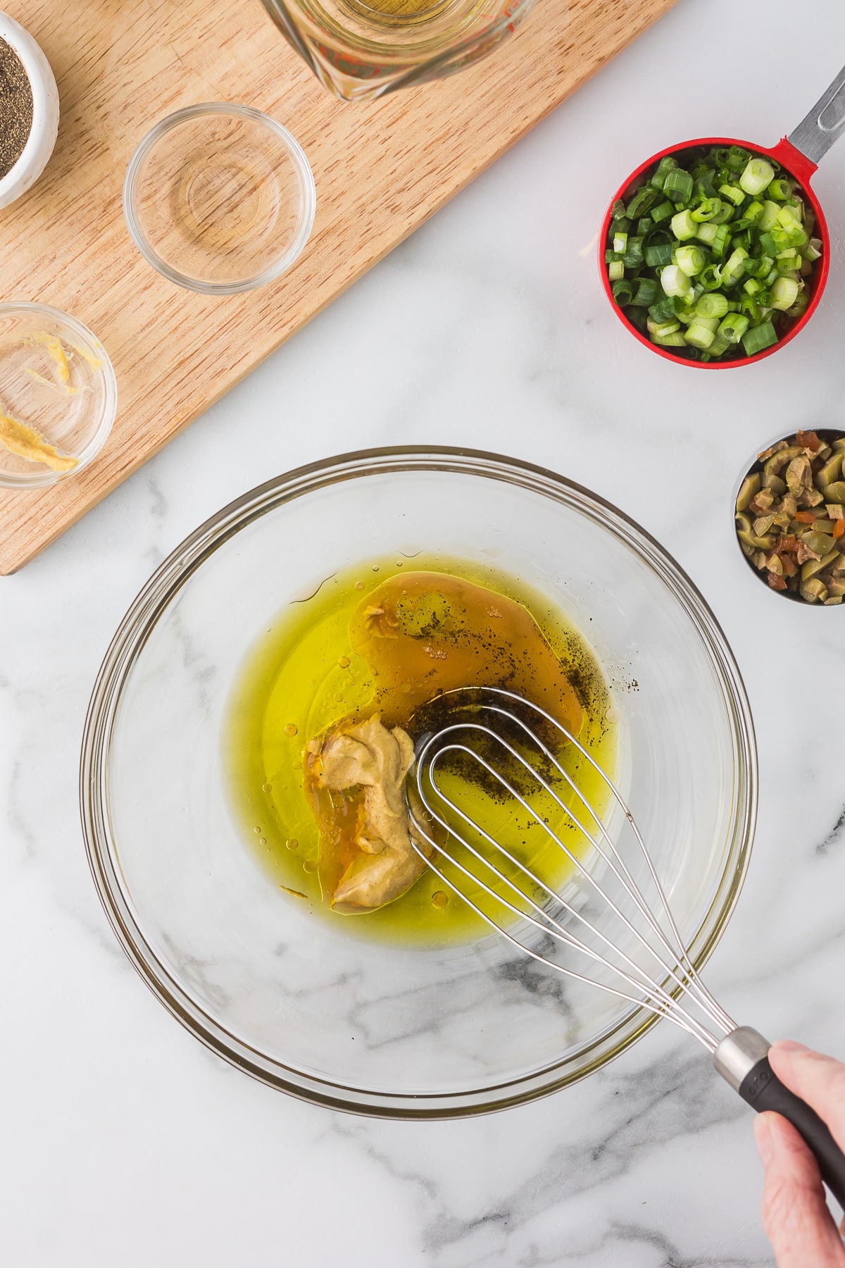 A glass bowl is filled with olive oil, mustard, and black pepper. A whisk is set inside the bowl, resting on the mix. Chopped green onions and olives are placed nearby on a marble countertop.