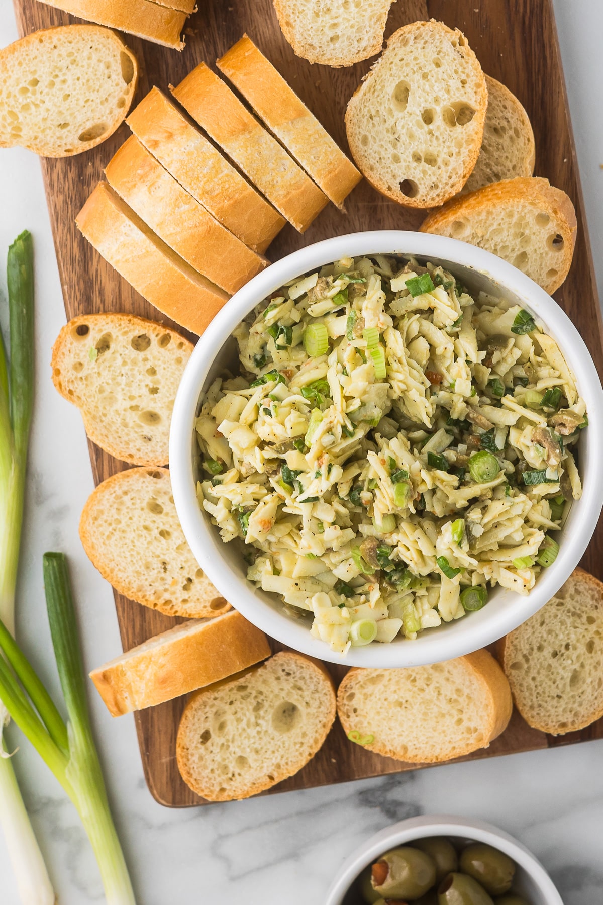 A bowl of pasta salad with orzo, green onions, and peas sits on a wooden board surrounded by slices of fresh baguette. Green onions lay alongside, and a small dish of green olives is visible in the corner.