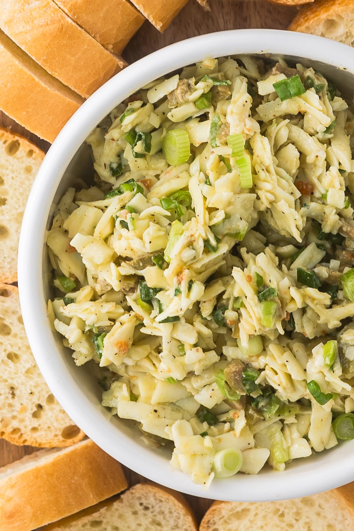 A white bowl filled with crab dip garnished with chopped green onions, surrounded by slices of baguette on a wooden surface.