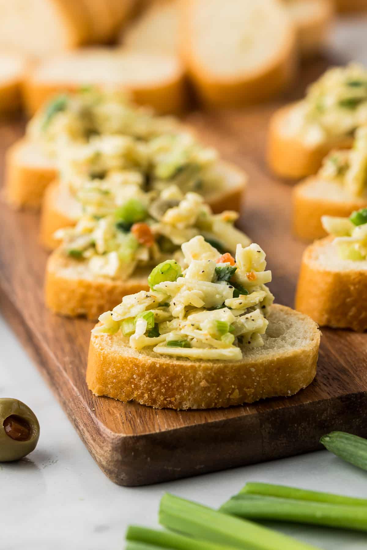 Slices of bread topped with a mixed salad featuring chopped vegetables and herbs are arranged on a wooden board. An olive and pieces of green onion are visible nearby on a light surface.