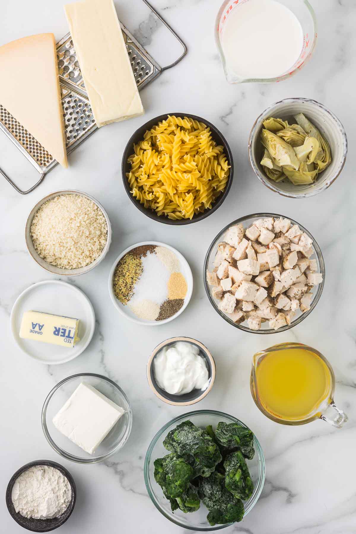 An assortment of ingredients on a marble surface, including grated cheese, fusilli pasta, artichoke hearts, spices, cooked chicken pieces, butter, sour cream, broth, frozen spinach, cream cheese, flour, and a small jug of milk.