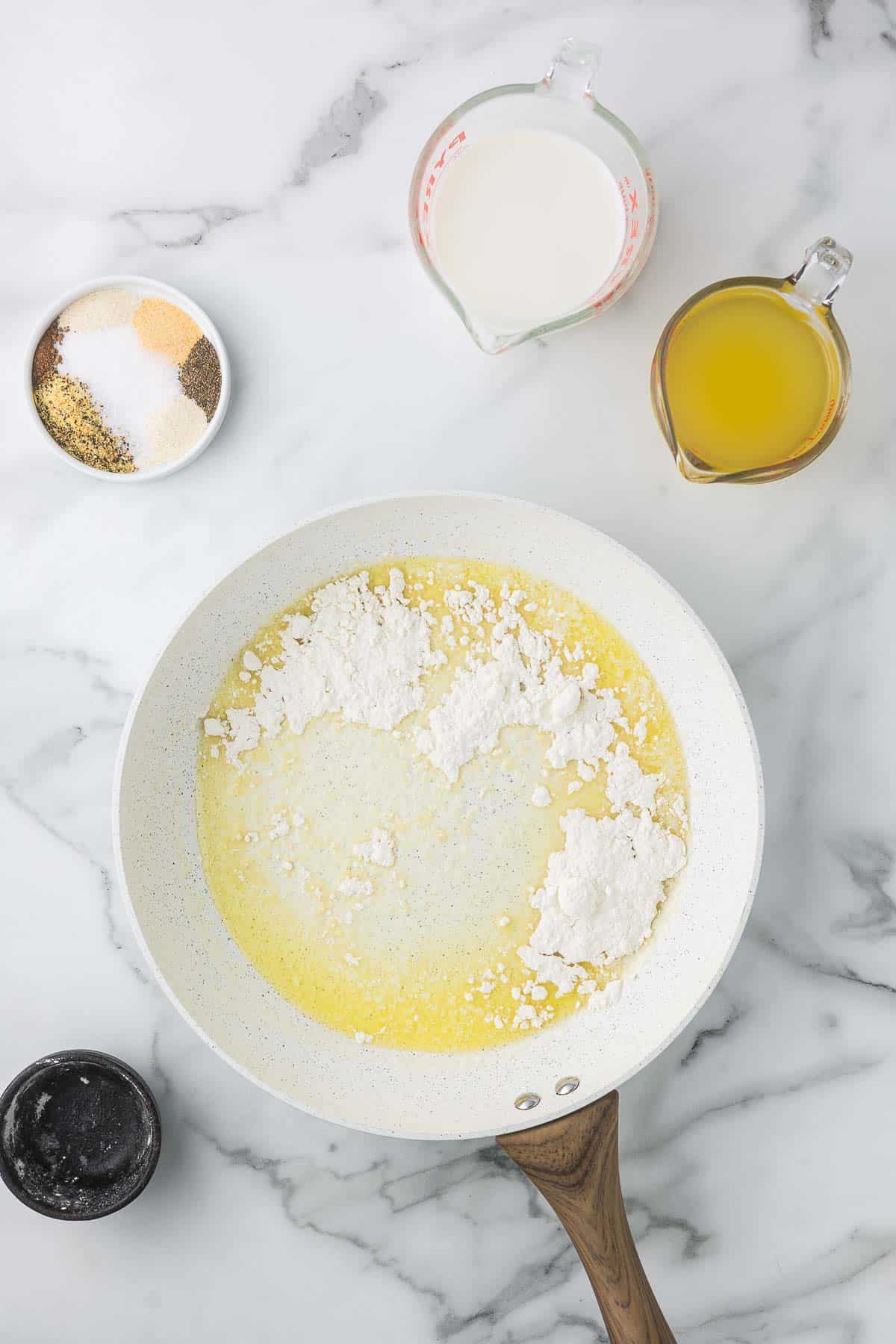 A white frying pan on a marble countertop contains melted butter and flour. 