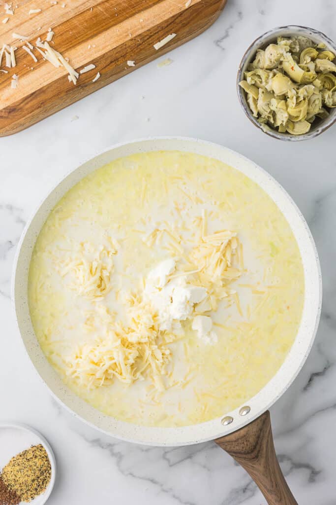 A white skillet with sauce, cream cheese, and white cheddar cheese melting before adding additional ingredients to make spinach artichoke chicken pasta.