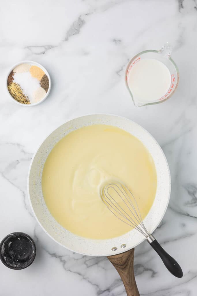 A skillet with creamy sauce being whisked on a marble surface, surrounded by small bowls of seasonings and a measuring cup of milk.