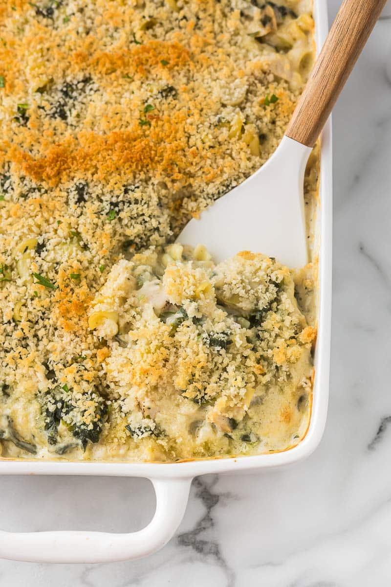 A casserole dish with spinach artichoke pasta in it and a spatula getting ready to serve.