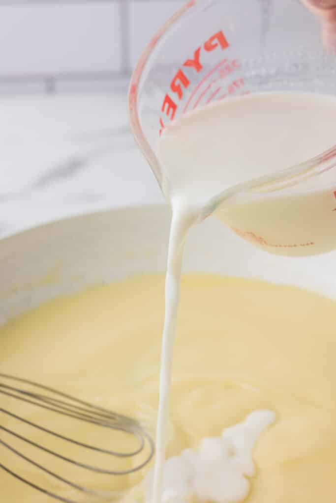 Pouring milk from a measuring cup into a saucepan, where a whisk rests in a yellow mixture. 