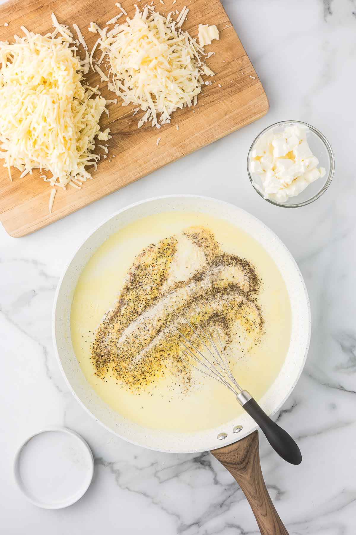 A pan on a marble countertop holds a whisk in creamy sauce sprinkled with black pepper and seasonings.