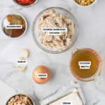 Ingredients for a dish are arranged on a marble surface: cooked shredded chicken, Rotel, frozen corn, seasonings, garlic, onion, chicken broth, great northern beans, and cream cheese.