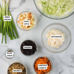 Top-down view of labeled ingredients for cabbage salad: green cabbage, green onions, diced apple, mayonnaise, craisins, celery seeds, shredded carrots, walnuts, white vinegar, and sugar, arranged on a marble surface with an apple on the side.