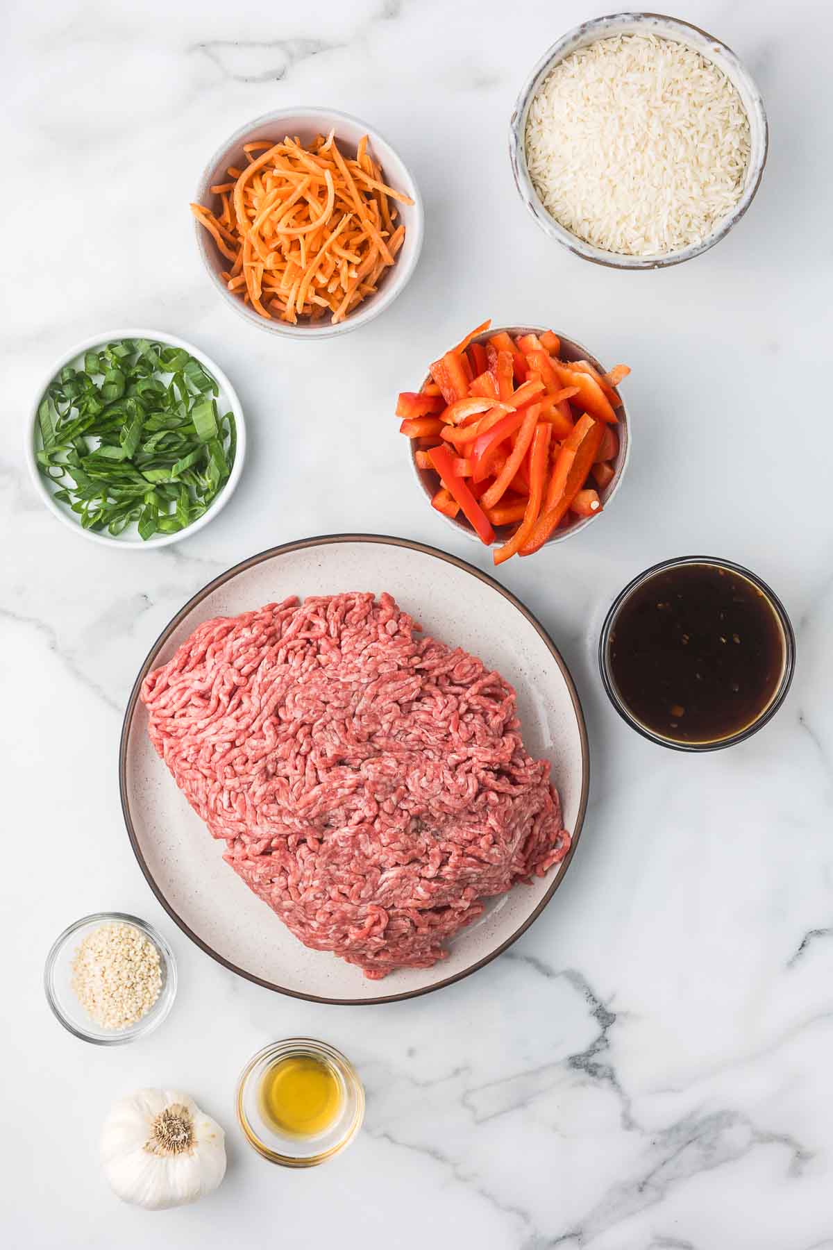 The ingredients to make teriyaki beef bowls including ground beef, teriyaki sauce, sesame seeds, garlic, green onions, red bell peppers, carrots and sesame oil.