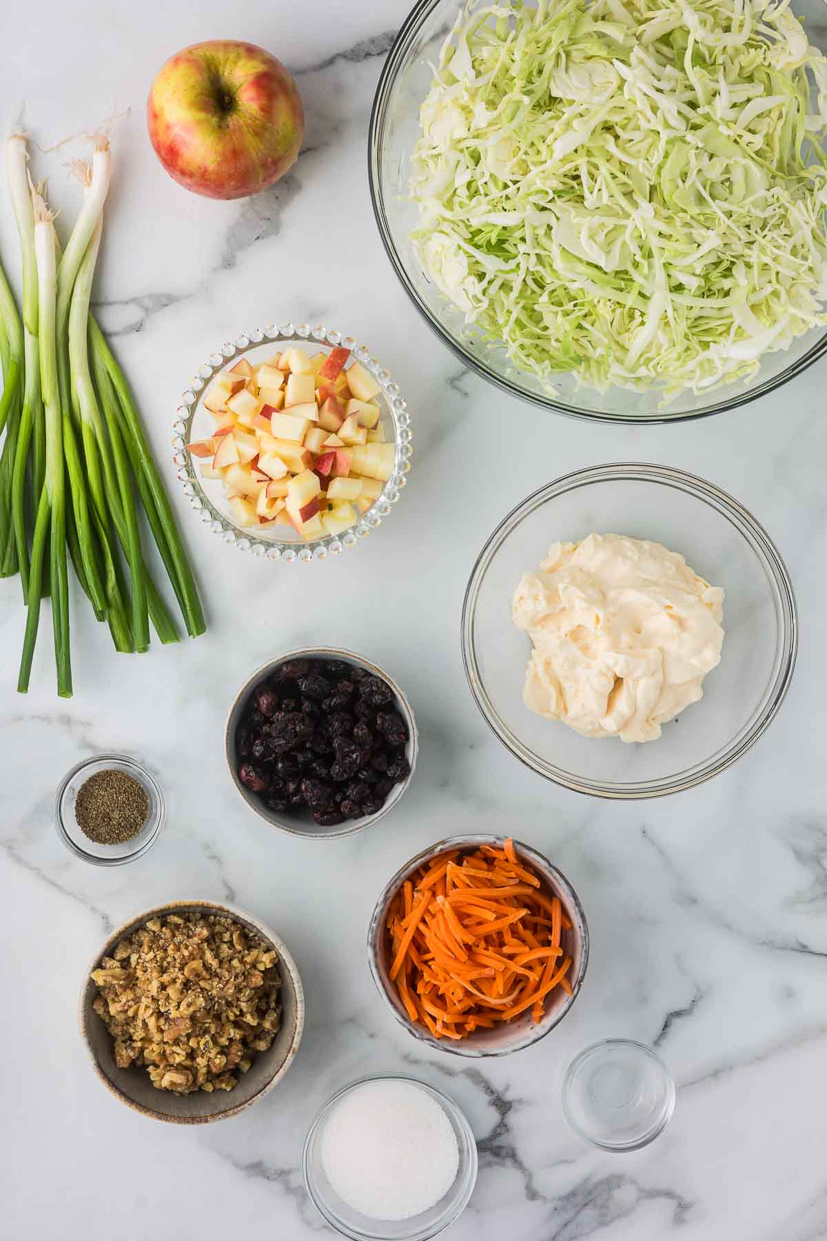 Ingredients for cabbage salad on a marble surface: chopped cabbage, diced apples, green onions, mayonnaise, raisins, julienned carrots, walnuts, black pepper, and sugar. Whole apple and green onions are also present.