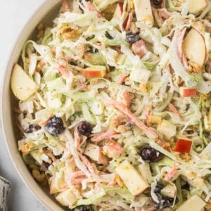 A bowl of coleslaw featuring shredded cabbage, sliced apples, carrot strips, walnuts, and dried cherries, all mixed in a creamy dressing.