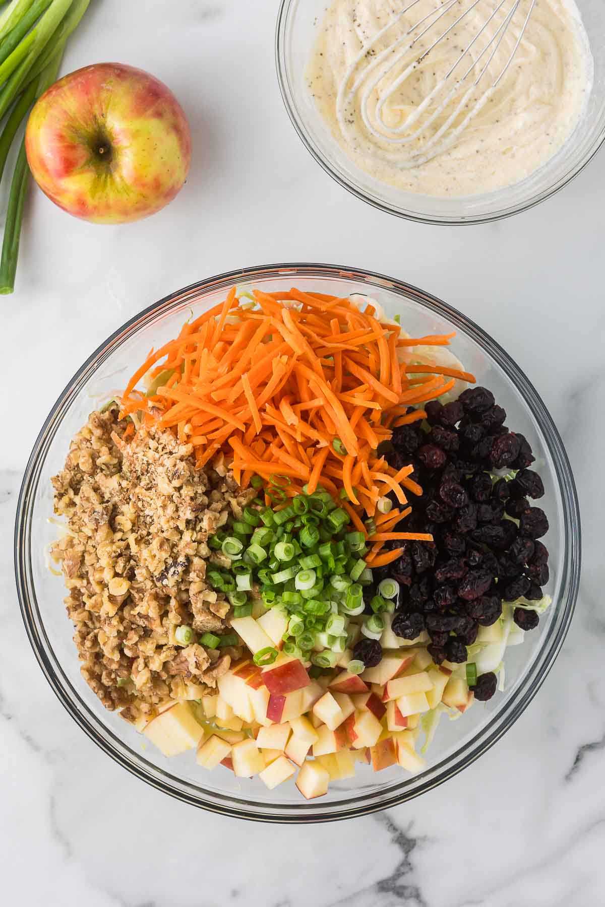 A glass bowl on a marble countertop contains shredded carrots, chopped walnuts, sliced green onions, dried cranberries, and diced apples. 