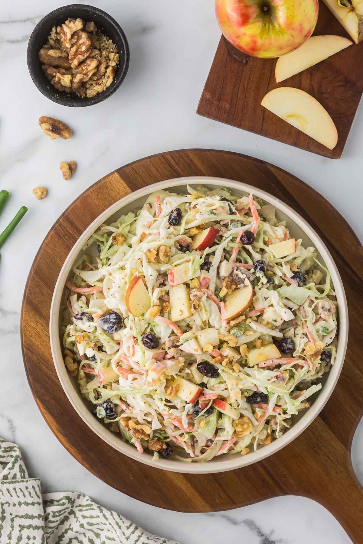 A bowl of coleslaw with sliced apples, walnuts, and raisins on a wooden board. 