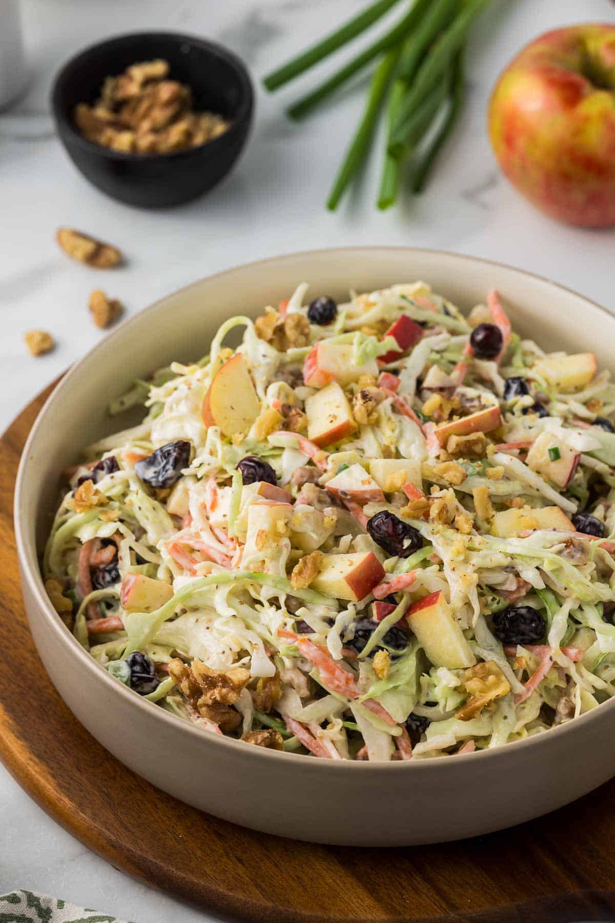 A bowl of coleslaw featuring shredded cabbage, apple slices, walnuts, and cranberries on a wooden board. Green onions, an apple, and walnuts are in the background.