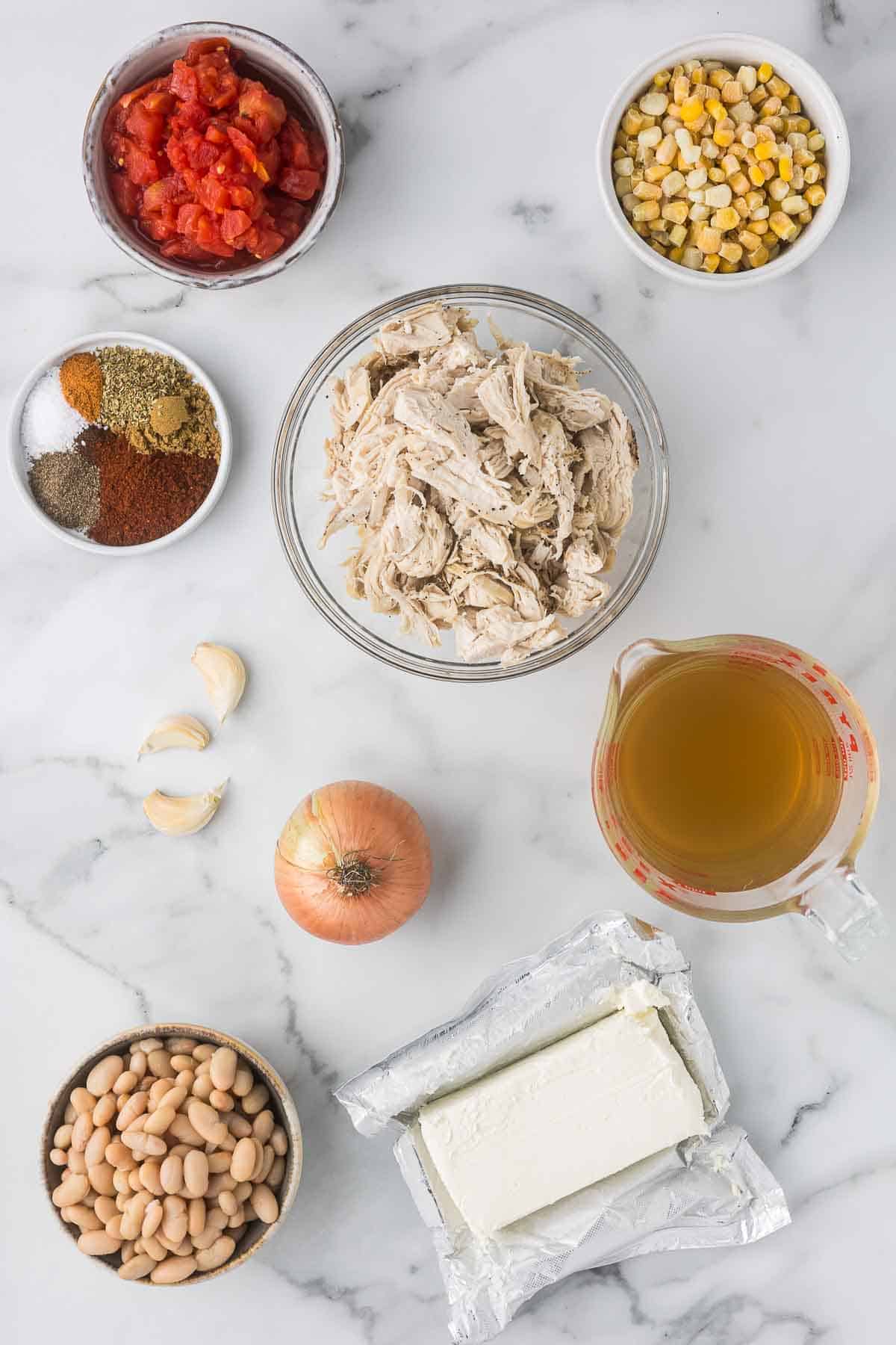 Ingredients for a dish arranged on a marble countertop, including shredded chicken, white beans, diced tomatoes, corn, chicken broth, cream cheese, spices, garlic cloves, and an onion.