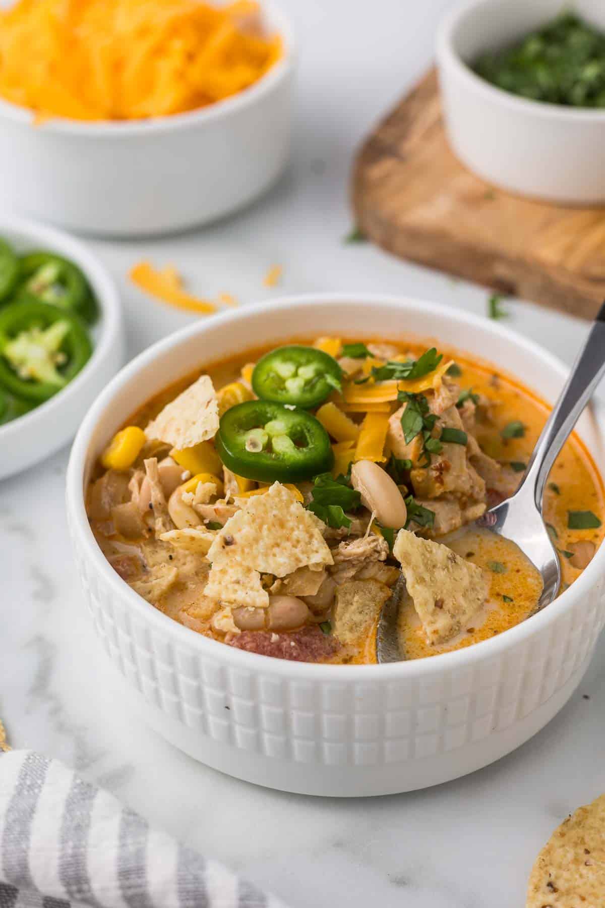 A bowl of creamy chicken chili topped with jalapeño slices, chopped herbs, and tortilla chips. It sits on a marble surface with a spoon, surrounded by bowls of shredded cheese, sliced jalapeños, and herbs in the background.