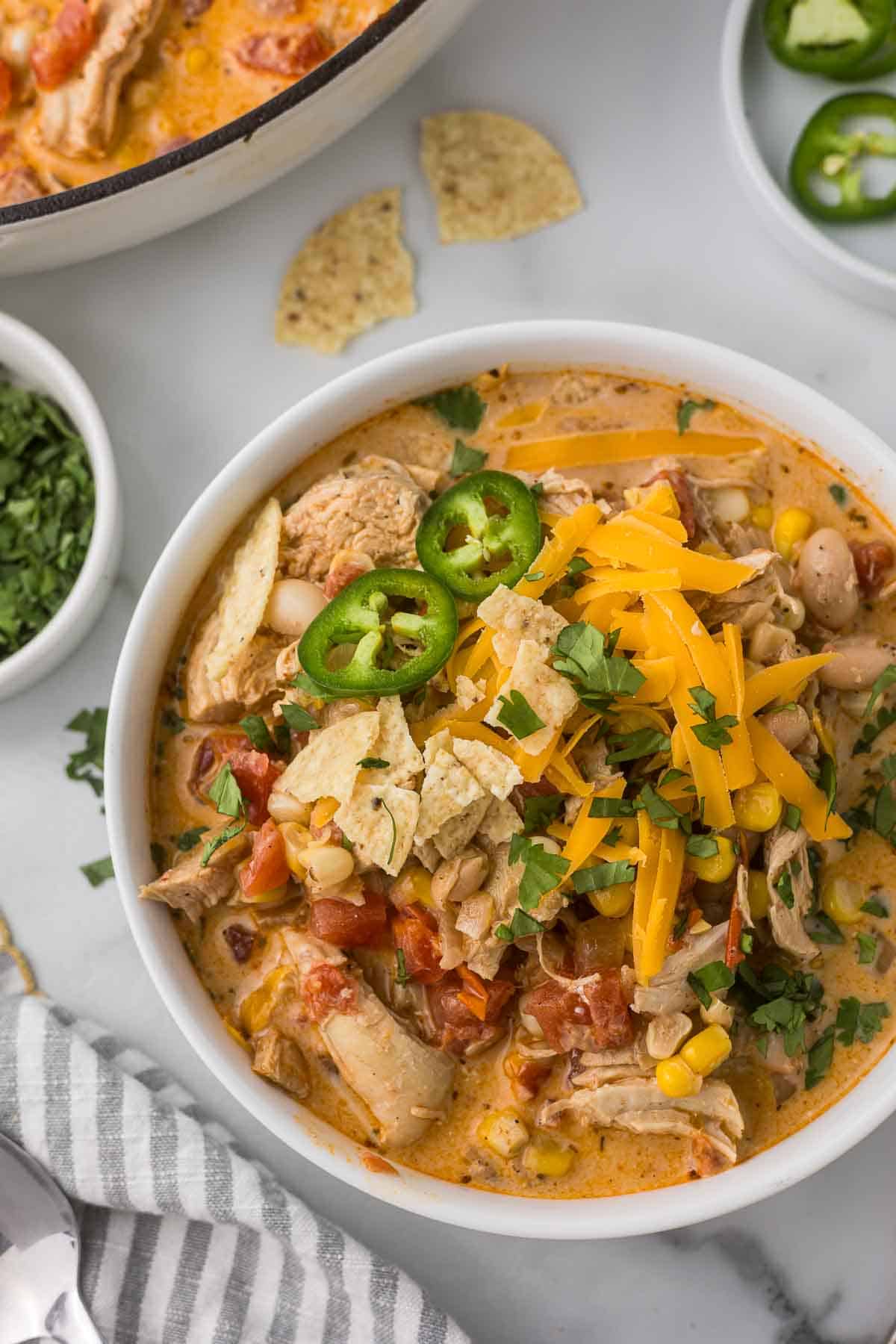 A bowl of chicken chili with cream cheese topped with shredded cheese, slices of jalapeño, tortilla chips, and chopped cilantro. The soup contains chunks of chicken, tomatoes, beans, and corn.