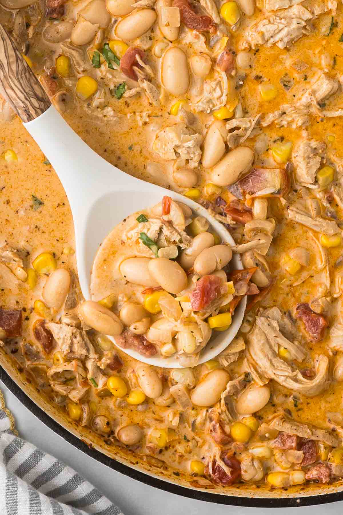 A creamy stew with white beans, shredded chicken, corn, and diced tomatoes being scooped with a white ladle. 