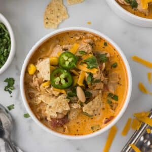 A bowl of creamy chicken soup with tortilla chips, sliced jalapeños, shredded cheddar, and cilantro on top. It also contains corn, beans, and tomatoes. There are shreds of cheese and chopped cilantro scattered nearby.