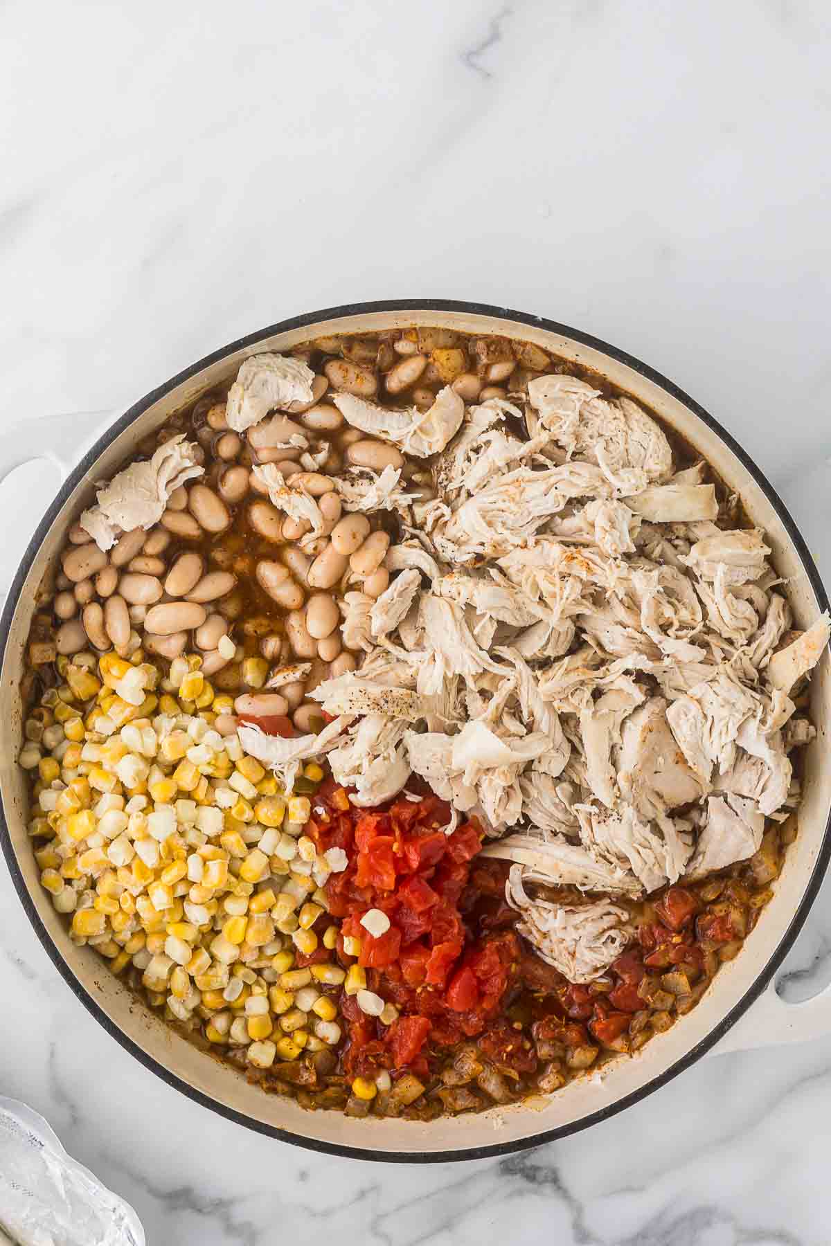 A pot filled with ingredients for a chili, including shredded chicken, white beans, corn, diced tomatoes, and spices. The mixture is arranged in sections on a white marble surface.