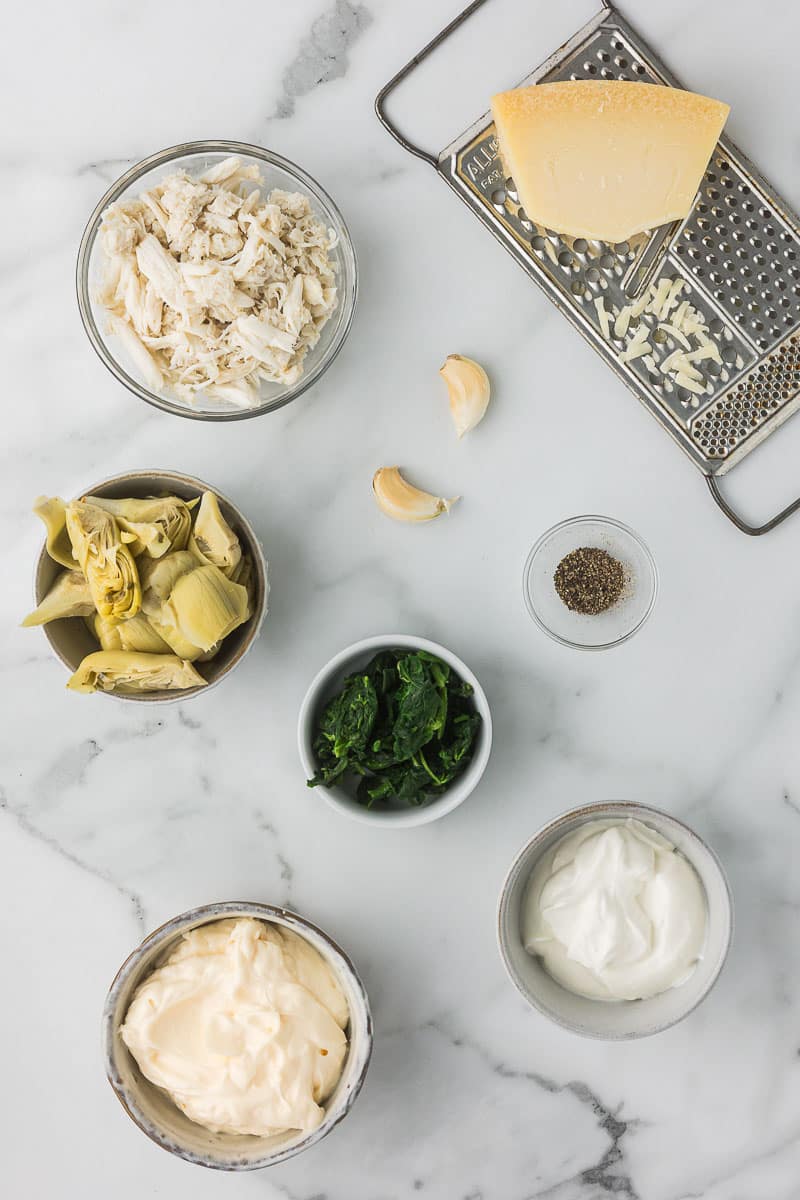 Ingredients for a recipe are laid out on a marble surface, including shredded cheese, garlic cloves, black pepper, artichoke hearts, spinach, mayonnaise, and sour cream.