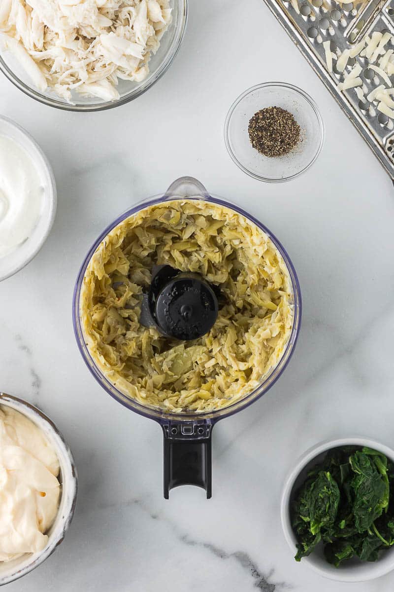 A food processor with chopped artichokes is surrounded by small bowls containing shredded cheese, crab meat, black pepper, mayonnaise, sour cream, and spinach on a marble countertop.