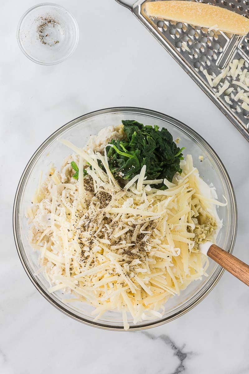 A glass bowl on a marble surface contains grated cheese, chopped spinach, rice, and black pepper. A wooden spoon rests inside. Nearby is a cheese grater and a small bowl of pepper.