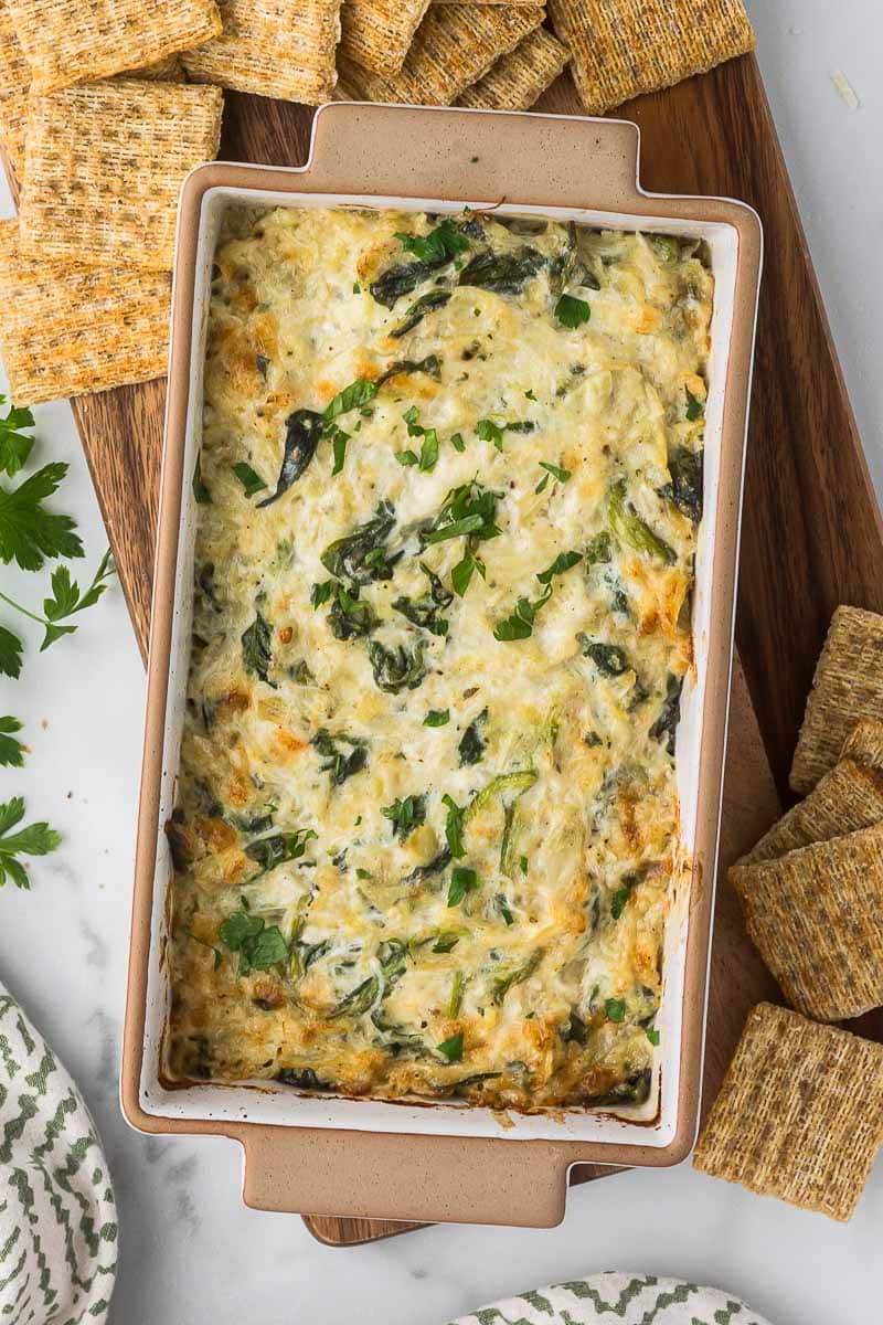 A rectangular baking dish filled with creamy spinach artichoke dip, garnished with chopped parsley.