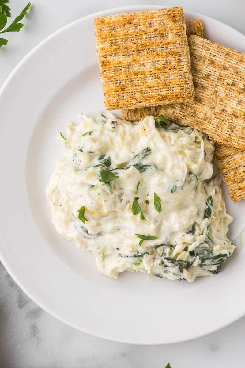 A white plate holds a serving of creamy spinach artichoke dip garnished with herbs, accompanied by several square whole-grain crackers. The dish is set on a marble surface with a glimpse of fresh green herbs nearby.