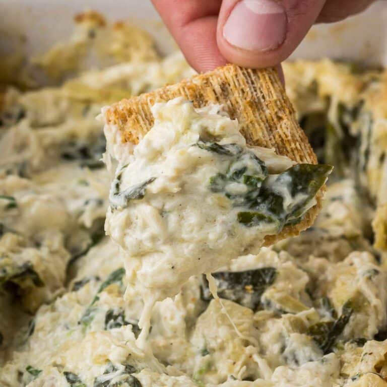 A hand holding a cracker dipped in creamy spinach artichoke dip, with visible spinach and artichoke pieces. The finger is slightly pressing on the cracker for grip. The background is blurred, focusing on the dip.