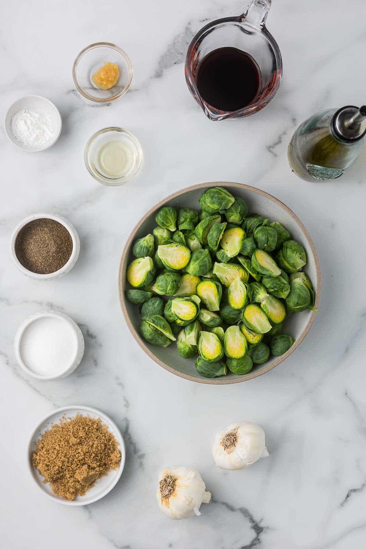 The ingredients to make teriyaki Brussels sprouts including a bowl of sprouts, salt and pepper, garlic, brown sugar, corn starch, rice wine vinegar, soy sauce, olive oil and ginger paste.